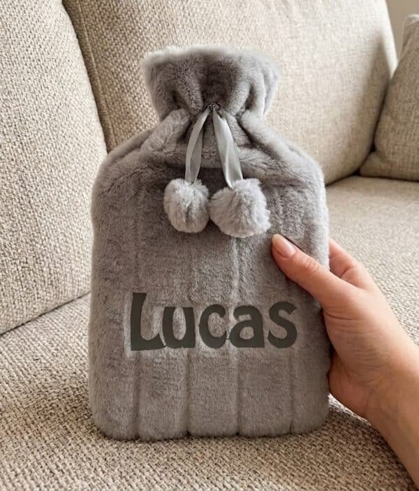 Grey fluffy personalised hot water bottle embroidered with Lucas, shown on a sofa
