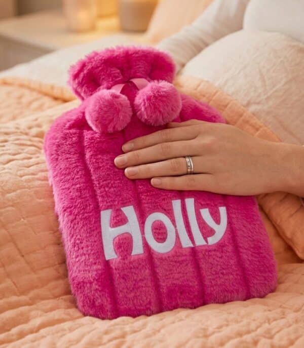 Pink fluffy personalised hot water bottle embroidered with Holly, shown on a bed