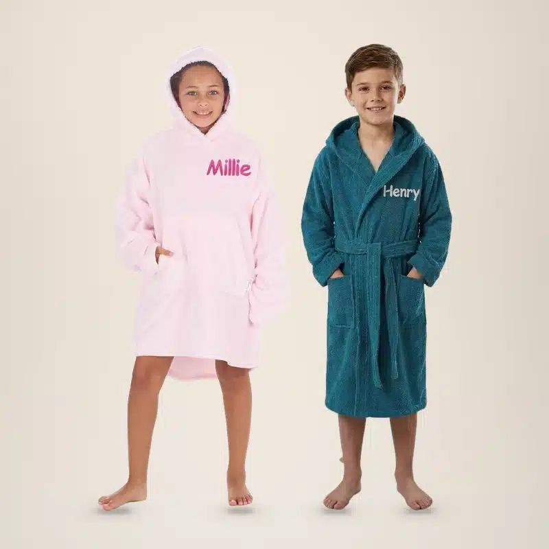 Girl in a pink personalised kids hoodie embroidered Millie next to a boy in a teal kids towelling robe embroidered Henry