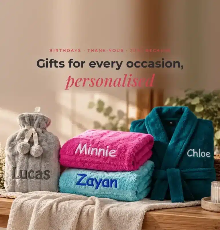 Personalised embroidered gifts: a grey Lucas hot water bottle, pink Minnie and aqua Zayan name towels and a teal Chloe robe on an oak bench