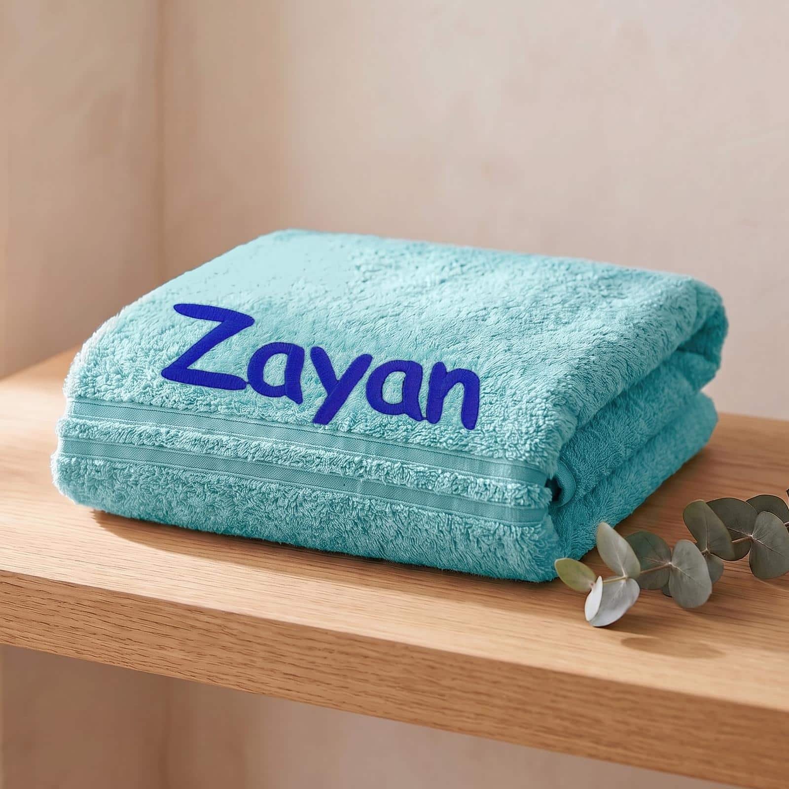 Folded aqua blue personalised towel with a name embroidered in navy, on a wooden shelf